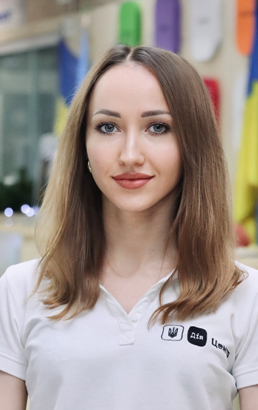 Жінка з довгим світло-русявим волоссям у білій футболці з логотипом на фоні табличок.